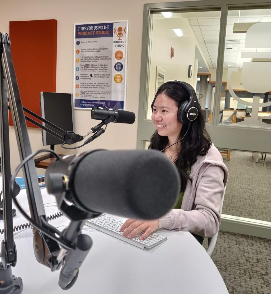 A person sitting at a keyboard, wearing headphones, in front of a microphone in the Podcast Studio.